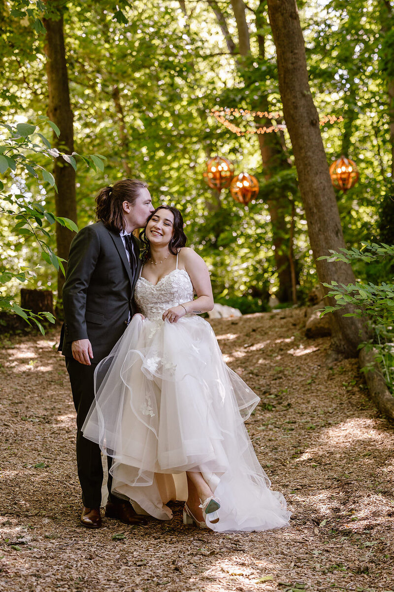 A bride and groom in the woods on their wedding day. They are standing next to one another. The groom is kissing the brides forehead, and the bride is smiling, and holding up her dress