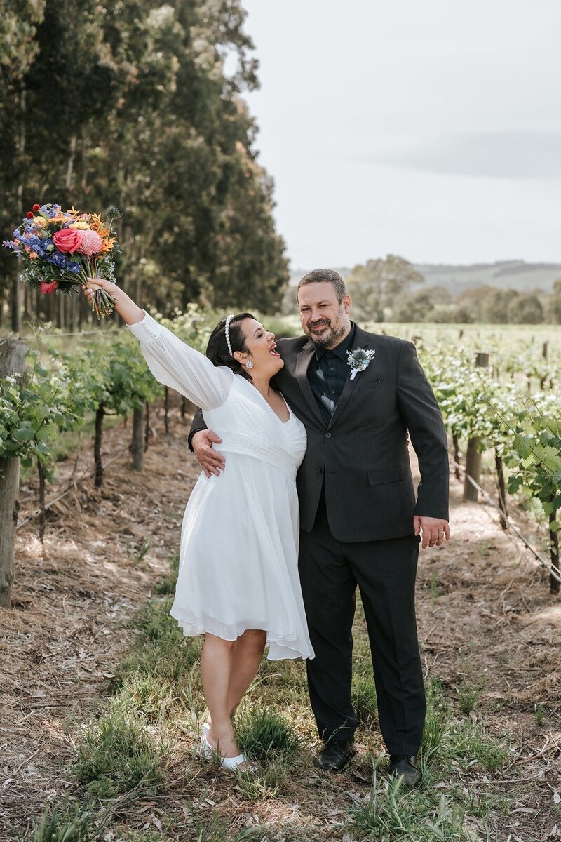 A happy bride holding a bright bouquet in the air hugging her new husand with a backdrop of vines