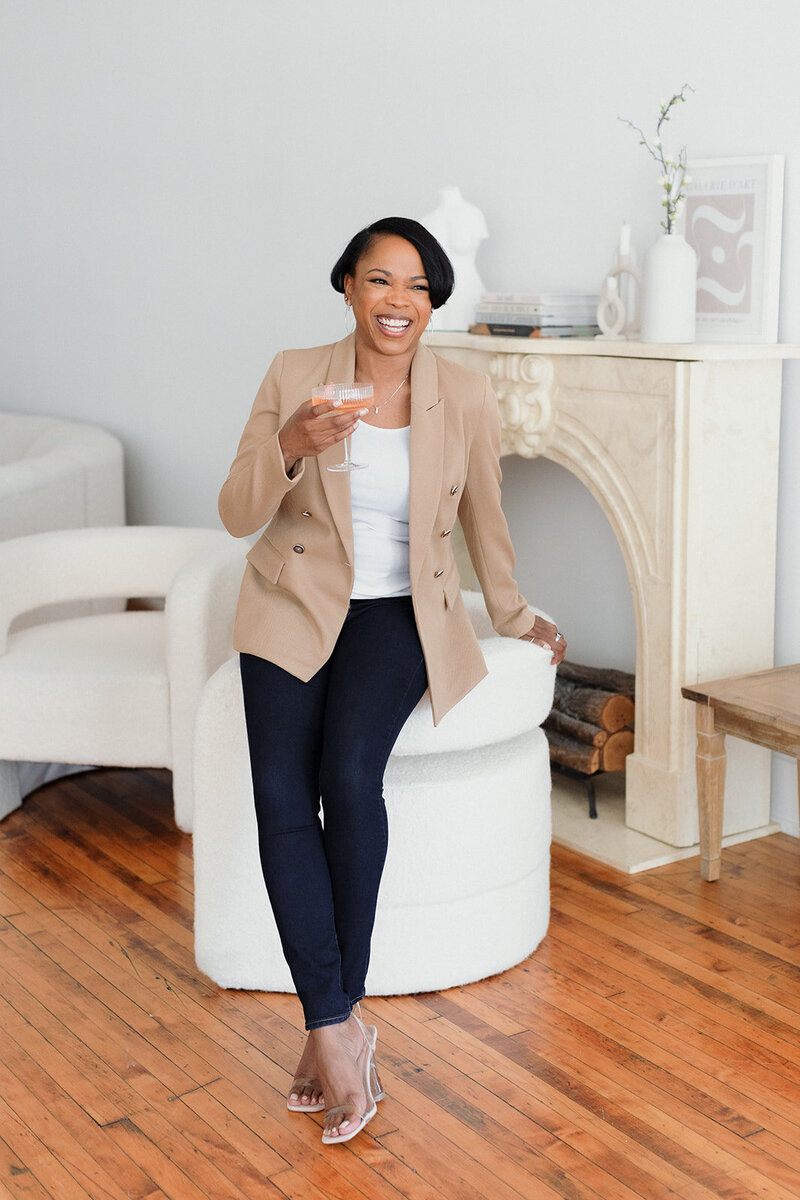 Beautiful woman with dark skin wearing jeans and tan blazer. She is holding a glass of champagne and laughing, while leaning on chair in front of white fireplace.