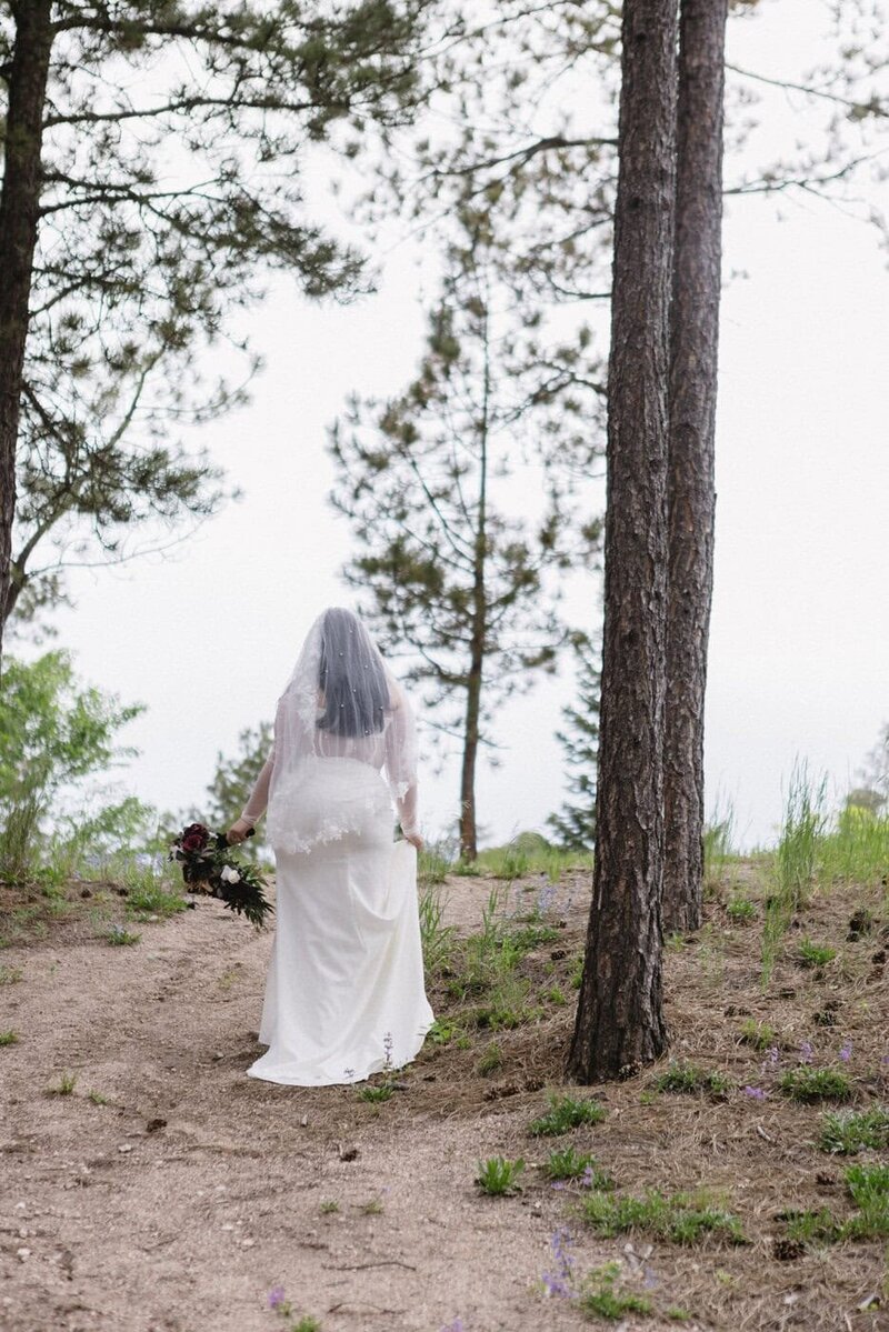 A view from behind of a bride walking up a hill with her bouquet and with tall trees next to her.