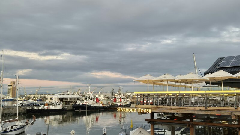 Exterior view of the Harbour House restaurant situated on the water at the V&A Waterfront in Cape Town, with boats docked nearby and a cloudy sky above.