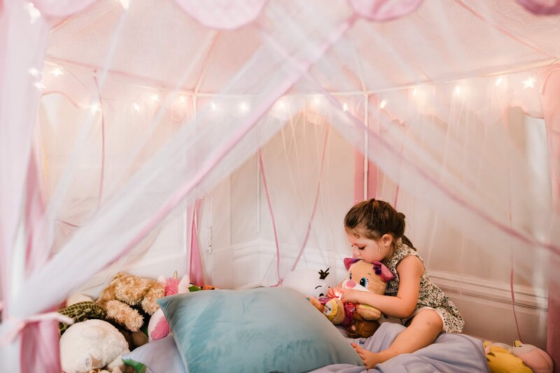 A child playing with stuffed toys in a cozy, tent-like canopy bed adorned with fairy lights, ready for photographing newborns at home.