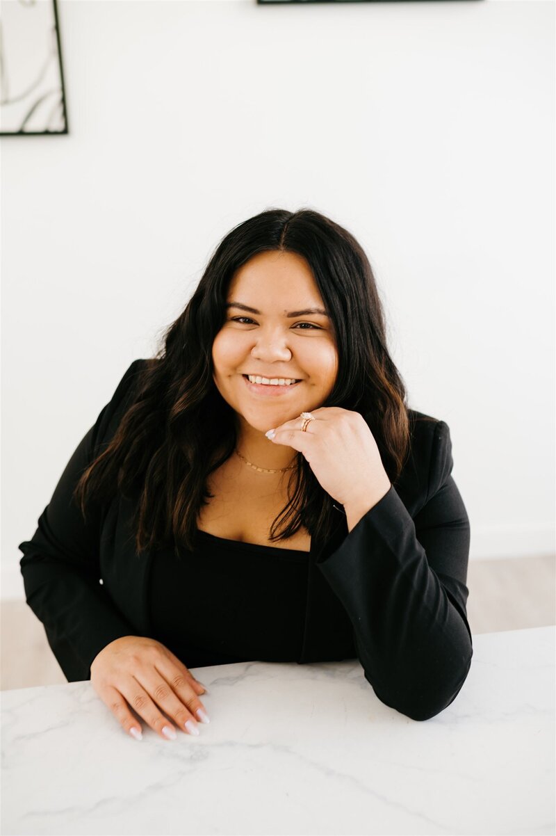 headshot of charlottesville wedding photographers  standing in a white studio and leaning one arm on a vintage green and gold chair while holding her camera in her hand