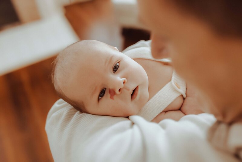 A close-up of a baby being held by an adult. The baby is looking up, wrapped in a white blanket, with a neutral expression. The adult's face is partially visible.