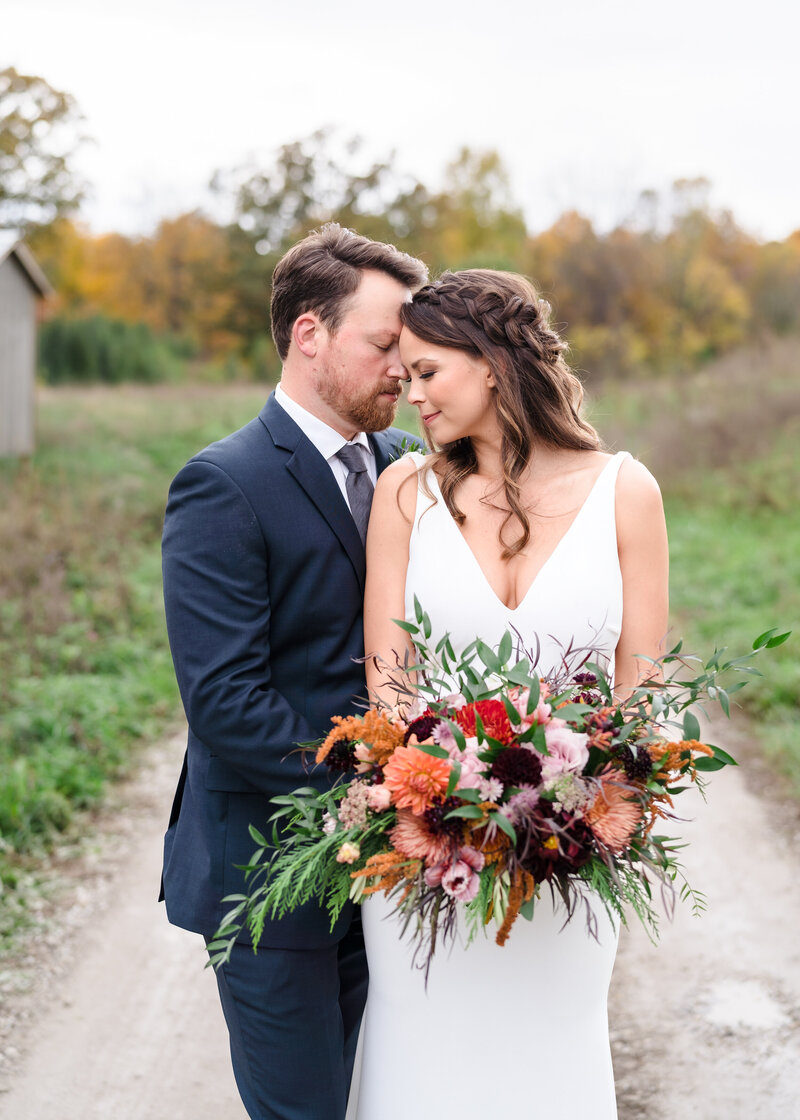 A bride holding a stunning boho bouquet  and surrounded by fall colors leans into her husband