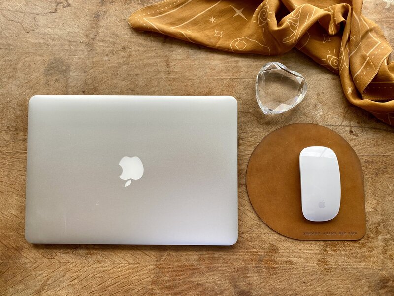 A flatlay with a macbook, apple mouse, leather mousepad, crystal heart and brown silk bandana.