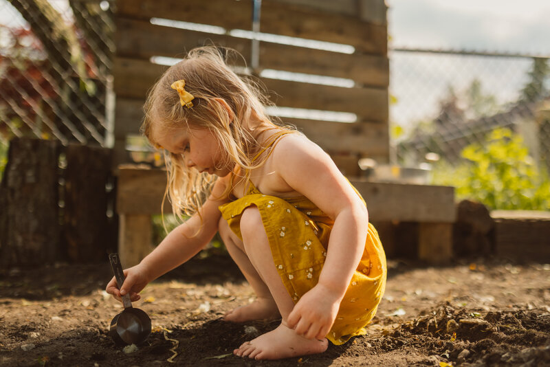 toddler girl wearing yellow dress with bow in her hair squats in dirt at homemade mud kitchen and uses measuring cup to scoop up a rock