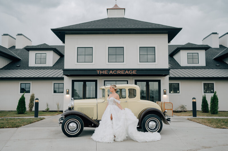 Bride in white off the shoulder wedding dress stands in front of a vintage yellow car at The Acreage Event Venue in Palmyra, NE - a venue near Lincoln NE. Photo by Anna Brace, a lincoln ne wedding photographer.