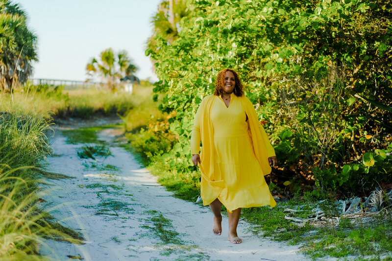 Certified life coach Claudine Martinell walking barefoot in a vibrant yellow dress amidst lush greenery, symbolizing her journey towards empowerment and personal growth.