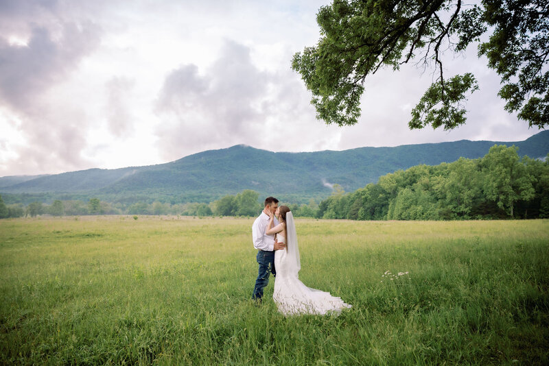 Cades Cove in the Smoky Mountains with bride and groom embracing in a meadow with the smoky mountains in the distance captured by gatlinburg photographers