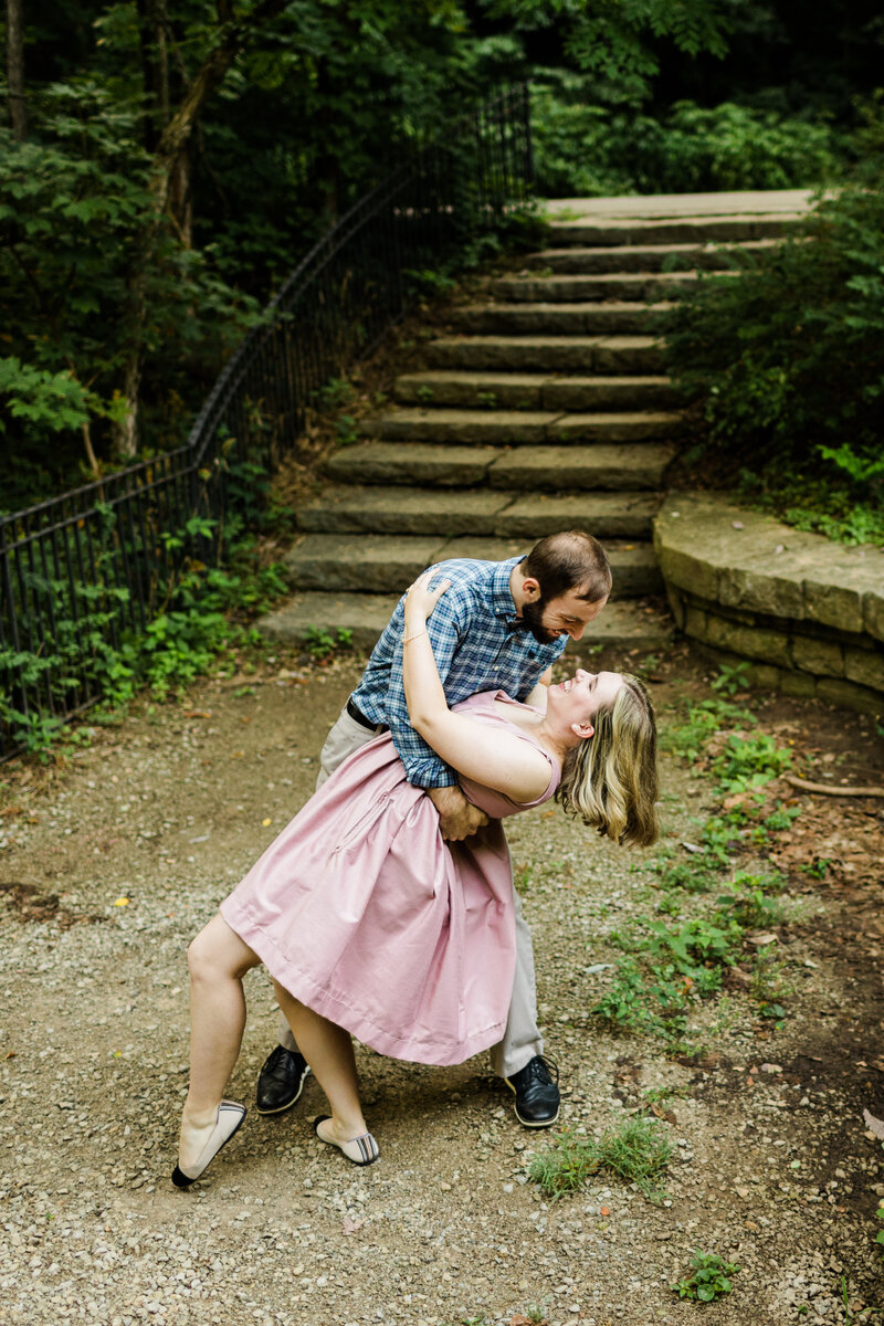An engaged man and wonam in a lush gravel park. The man is dipping the woman in a posed dance move.