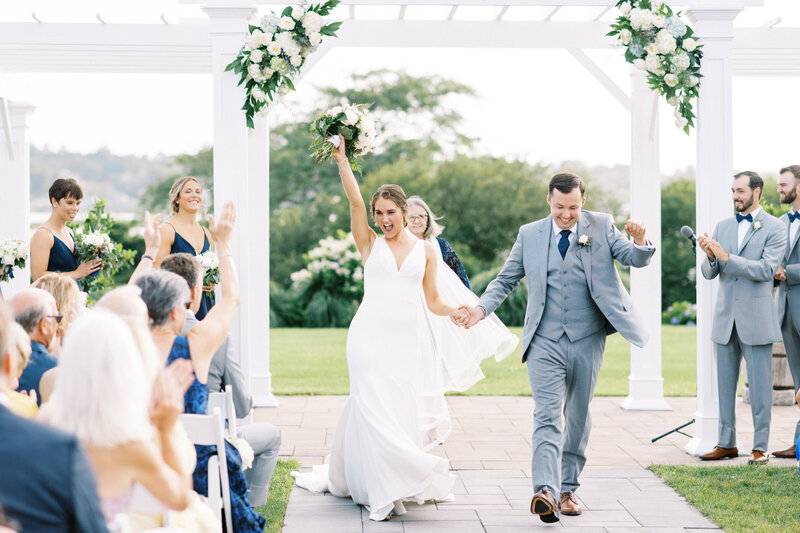 bride raises bouquet in excitement as she walks down aisle with her husband at Newport Wedding