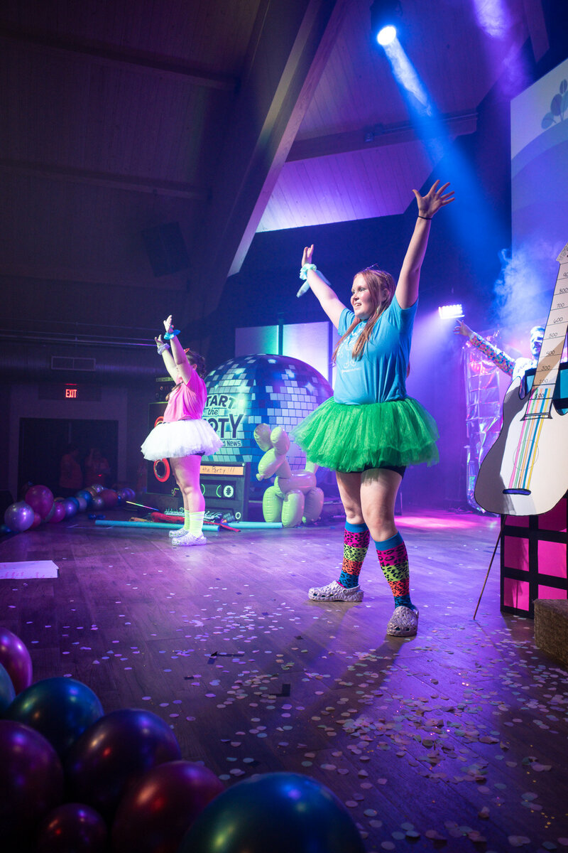 Two girls standing on stage in tutus with hands in the air.