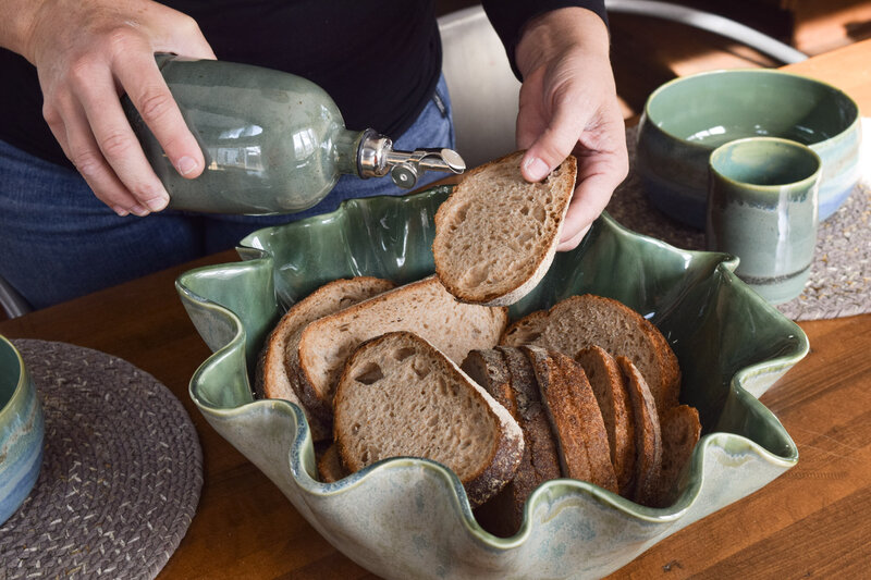 Artist pours oil onto dark sourdough bread being served in a sage green handmade ceramic wave serving bowl
