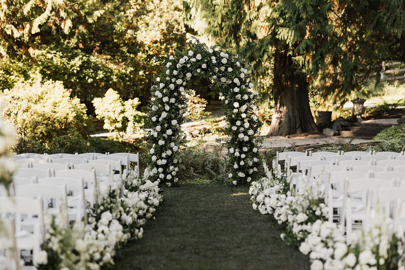 Flower-adorned archway surrounded by white chairs in an outdoor ceremony setting