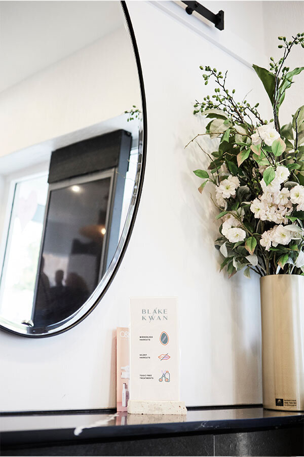 Modern salon interior at Blake & Kwan, featuring an oval mirror, floral arrangements, and branding elements for an inclusive space.