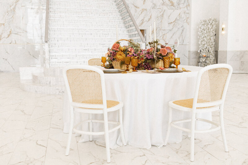 White-linen round table with amber glassware and an autumn centerpiece, surrounded by white cane-back chairs in a bright venue.