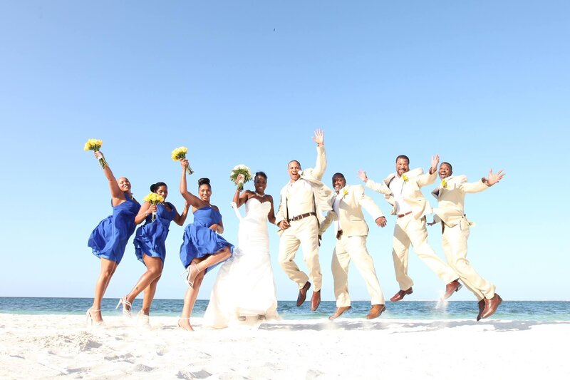 A bride, groom, and wedding party joyfully jump in the air on a sandy beach, celebrating their destination wedding in Mexico.