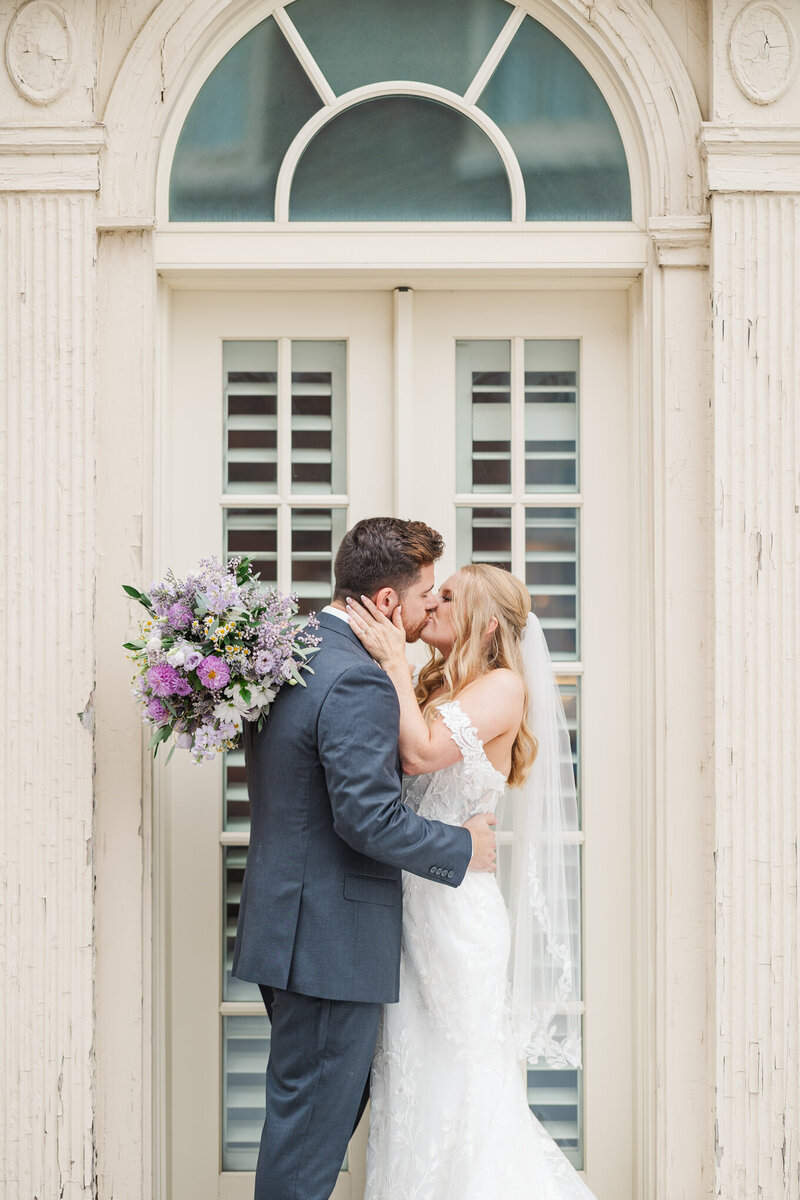 The happy couple kisses in front of the iconic balcony doors of Omaha's Magnolia Hotel. Natural light wedding photography.