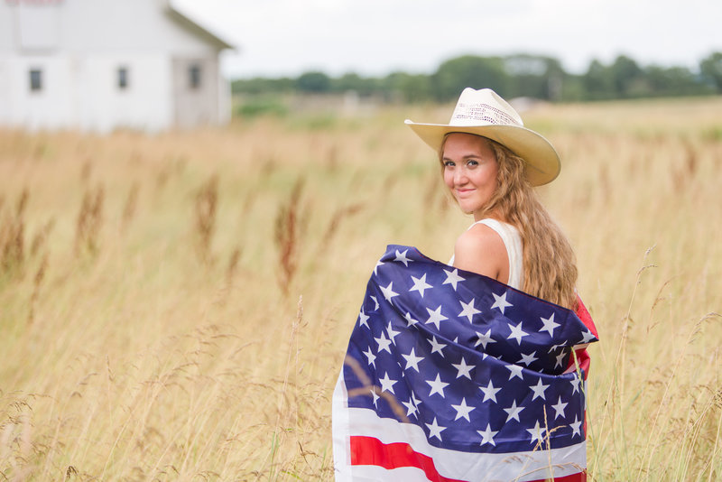 waynedale high school senior girl standing in wheat field with american flag wrapped around her with cowgirl hat on looking back over her shoulder towards camera photographed in wayne county ohio by jamie lynette photography canton senior photographer