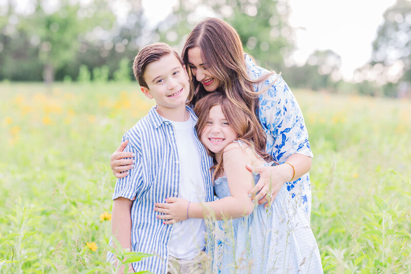 Mother hugging her son and daughter while smiling outdoors in a grassy field in Southeast Michigan.