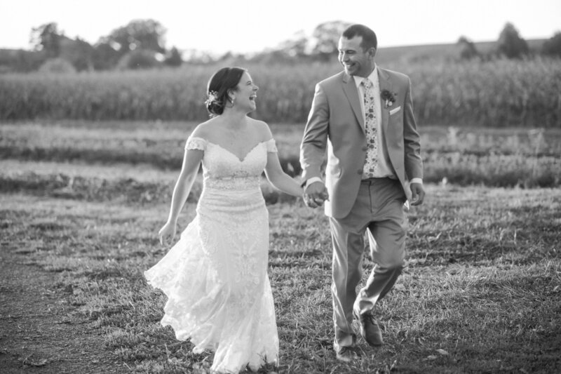 A bride in a lace wedding dress and a groom in a suit walk hand in hand through a field during golden hour.