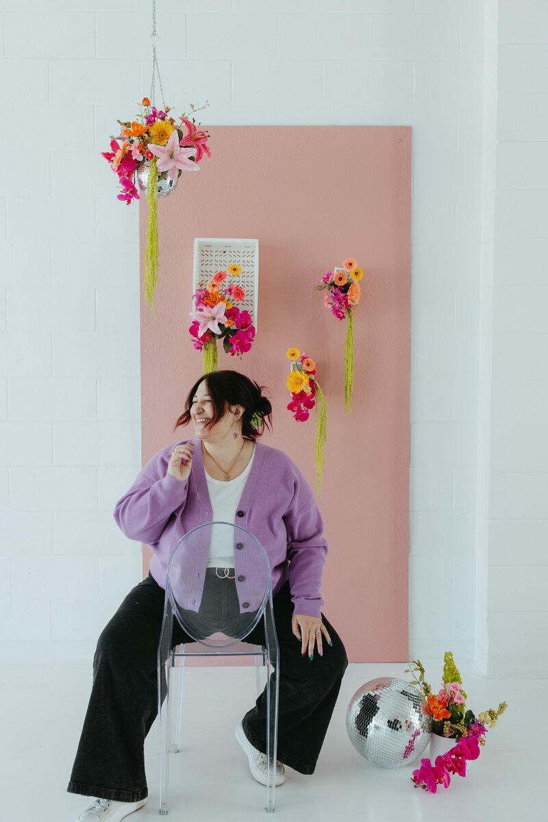 A person with dark purple hair styled in a loose updo sits on a clear acrylic chair, laughing and looking to the side. They wear a lilac cardigan over a white top with black pants and white sneakers. The background features a pink wall decorated with vibrant floral arrangements in pink, orange, and yellow, along with a hanging disco ball planter and a reflective disco ball on the floor.