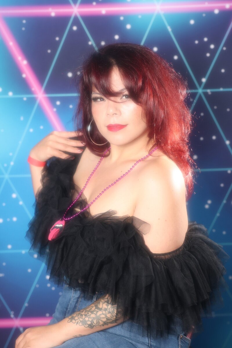 80's Glamour Shot Photo Booth featuring woman wearing black top posing in front of 80's themed blue and pink backdrop