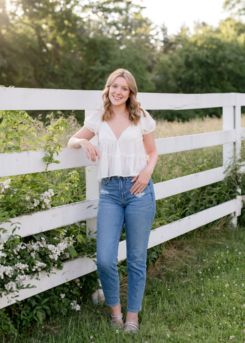 girl standing in front of a white fence in a field wearing a white shirt and jeans