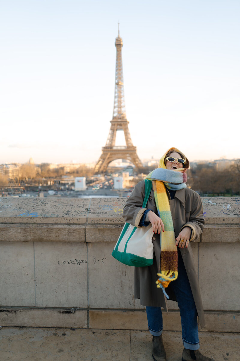 Bailee wearing a long coat and yellow sunglasses with a colorful scarf wrapped around her neck and head with the Eiffel Tower in the background
