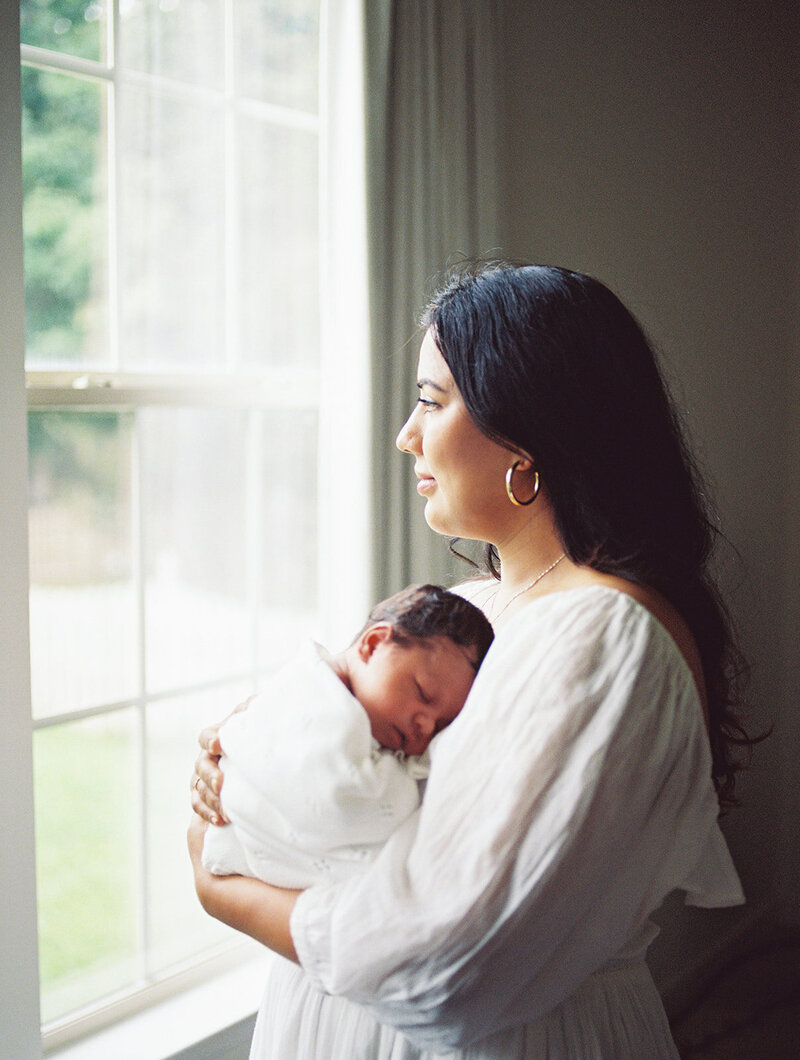 A mother holding her newborn and looking out of a window by Katie Stansfield Photography, a Richmond newborn photographer.