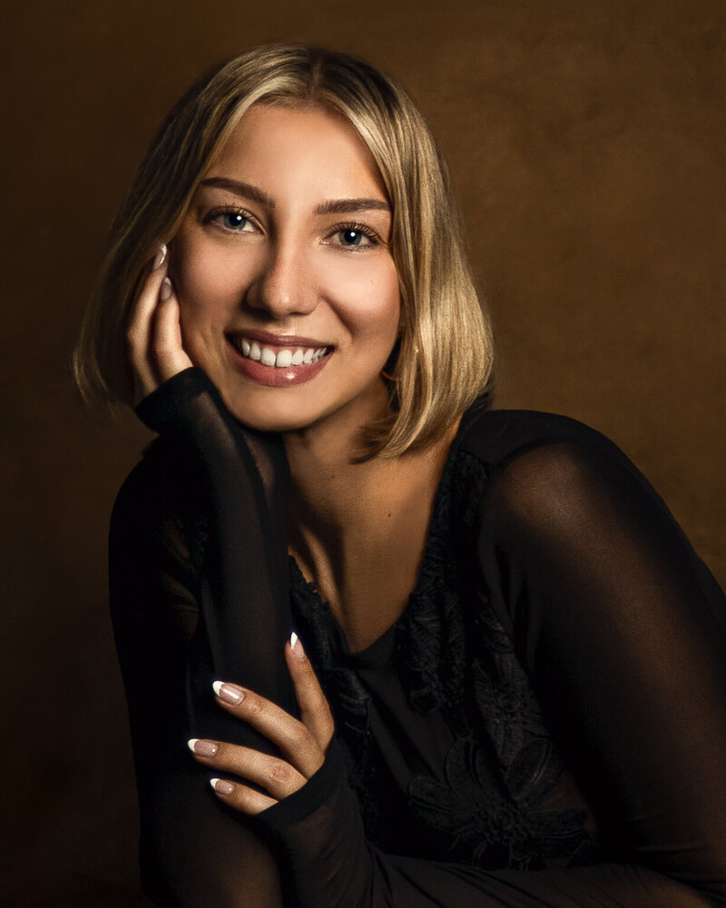 Smiling young woman  with hands in frame wearing sheer black top with lace on custom brown backdrop