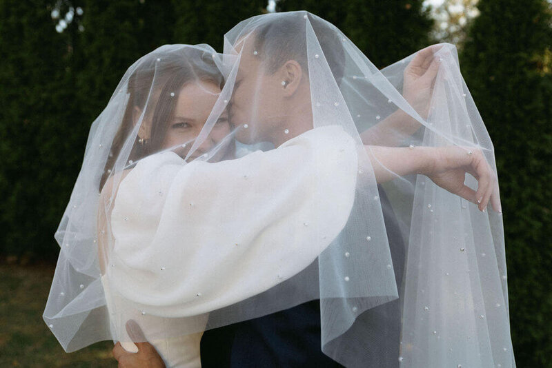 Couple in wedding attire embracing under a veil with pearl detials. Bride looks towards the camera while groom kisses her head