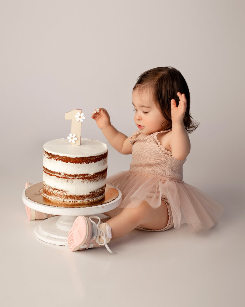 Stamford Cake Smash photographer captures one year old baby girl with long brown hair wearing a pale pink tutu dress with pink nike sneakers looking at her tall neutral birthday cake with frosting on it in front of a cream backdrop