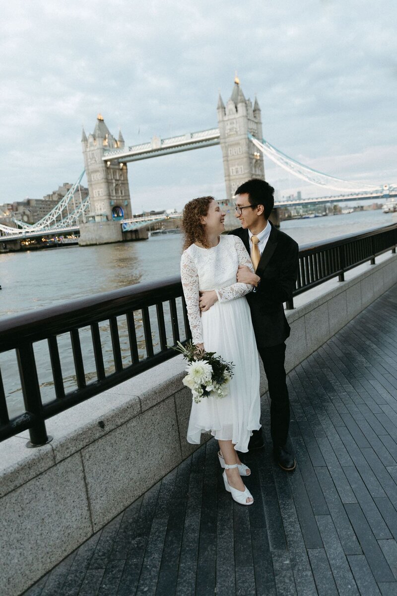 A couple smiles at each other in front of the Tower Bridge in a London elopement wedding.