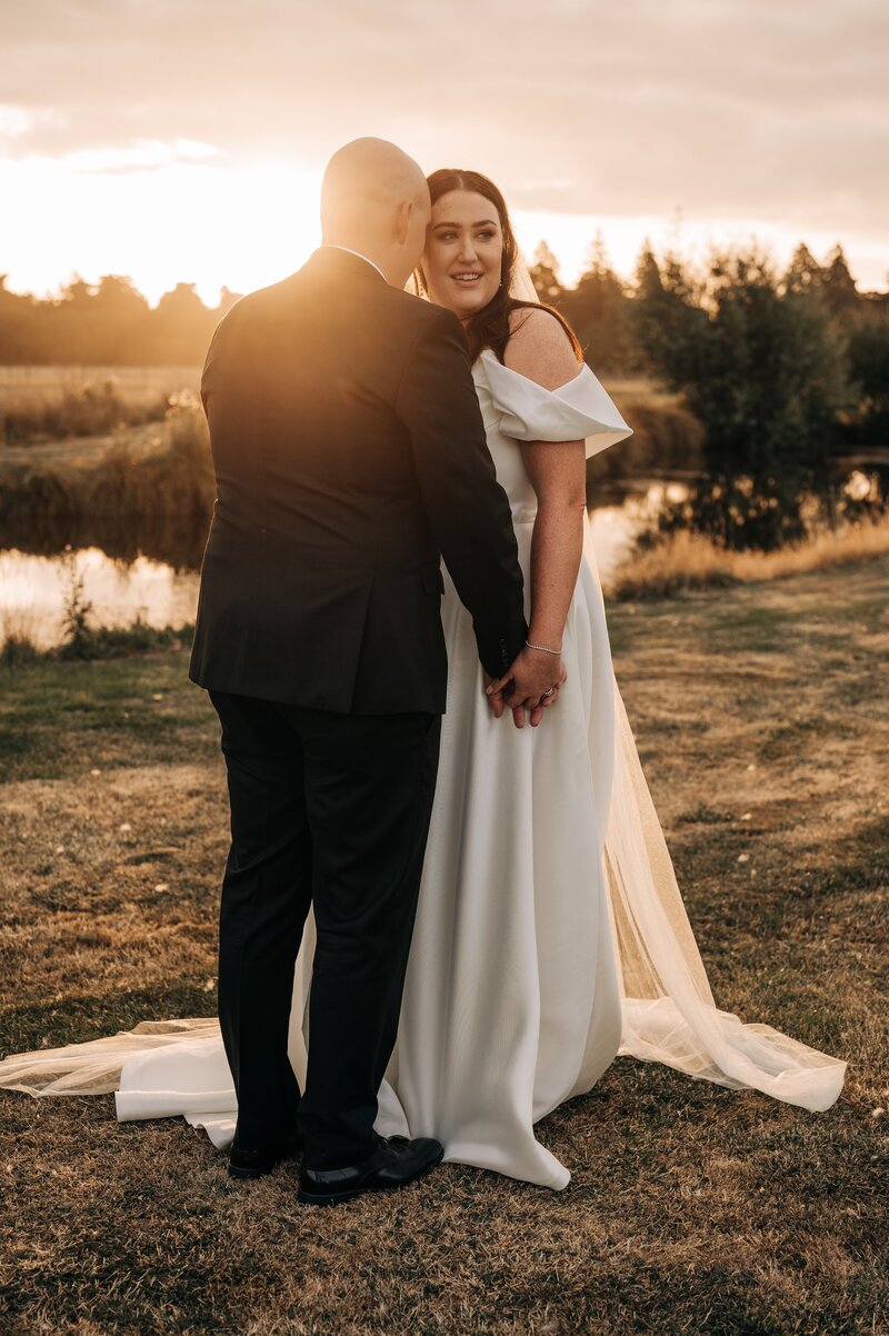 bride and groom stand together in front of pond at golden sunset on their wedding day at bangor farm in darfield