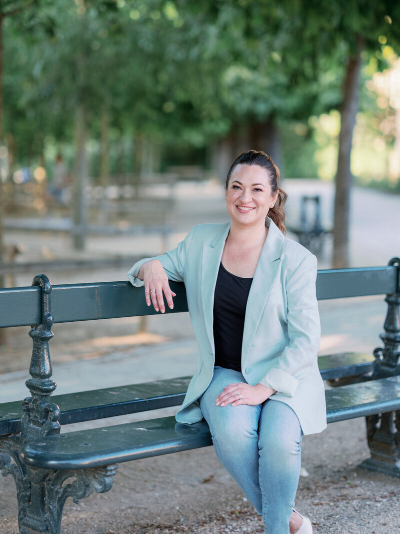 a woman in a blue blazer sitting on a park bench in paris smiling at the camera