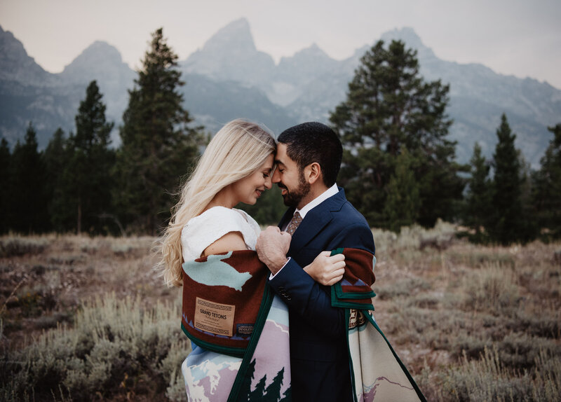 jackson hole wedding  with bride and groom embracing with a wool blanket wrapped around them as they stand close to one another and hold their heads together