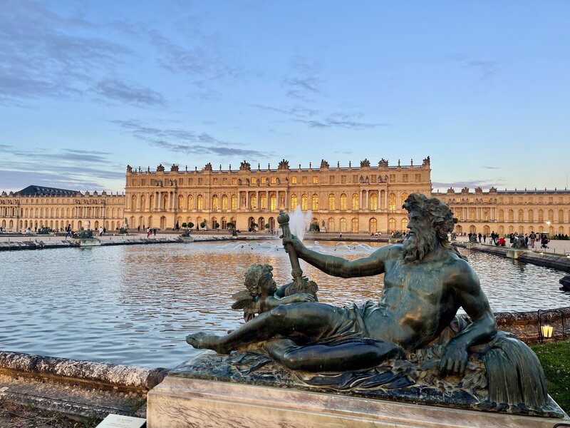 The Château of Versailles with ornate fountains and a classical statue in the foreground, showcasing its opulent gardens and grandeur.