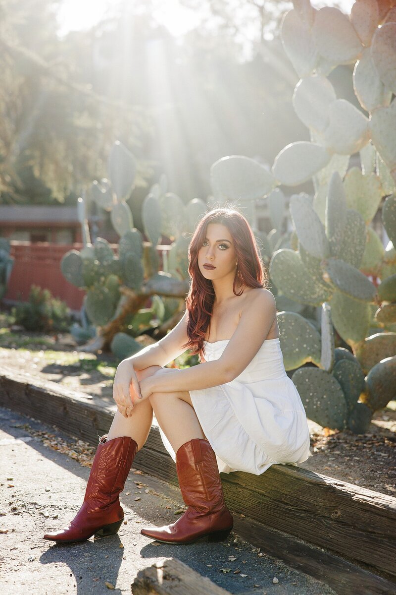 Boho senior girl in white dress and red cowgirl boots sitting by a cactus garden in Santa Clarita with golden backlight