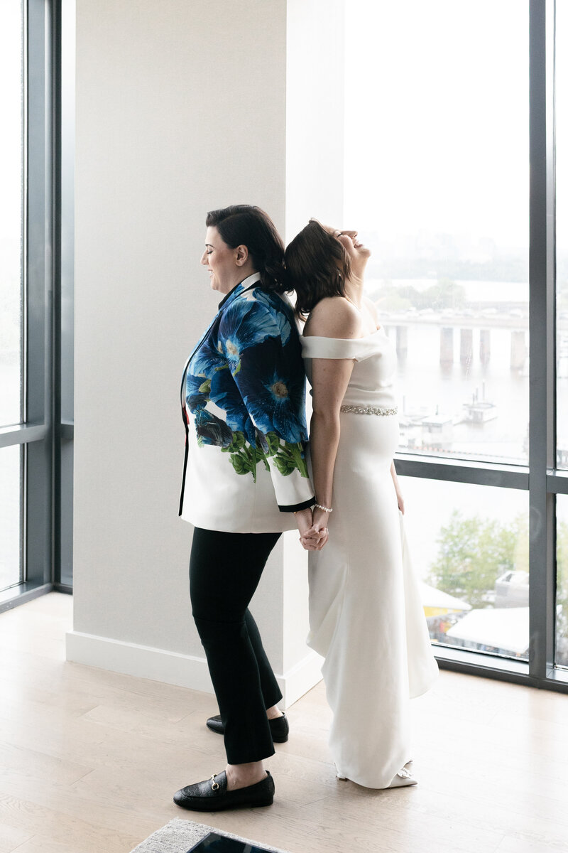 lesbian couple back to back holding hands smiling and laughing preparing for their wedding 1st look