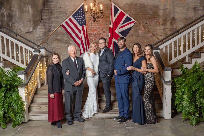 Seven people dressed formally stand on stairs decorated with string lights and ferns, with American and British flags behind them. The group smiles for the photo in front of a brick wall under a chandelier.