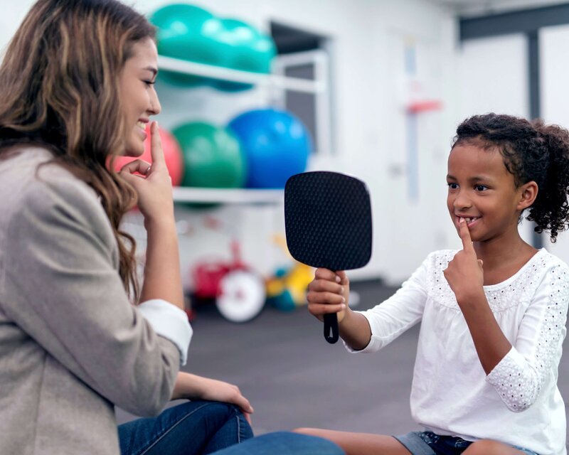 Speech-language pathologist using a mirror and letter chart to guide a young boy through articulation exercises during a speech therapy session in a bright, welcoming office.