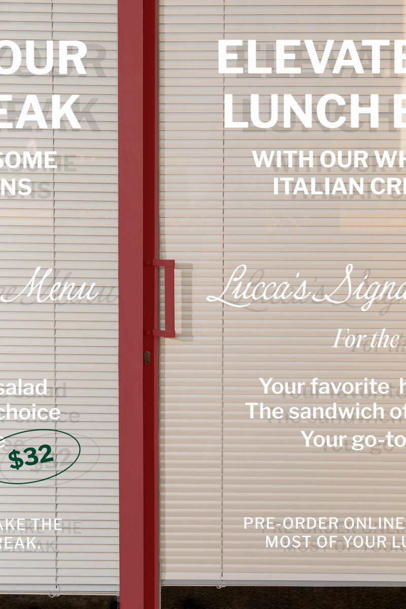 LUCCA storefront window with white vinyl lettering showcasing lunch menu and Italian sandwich shop branding.