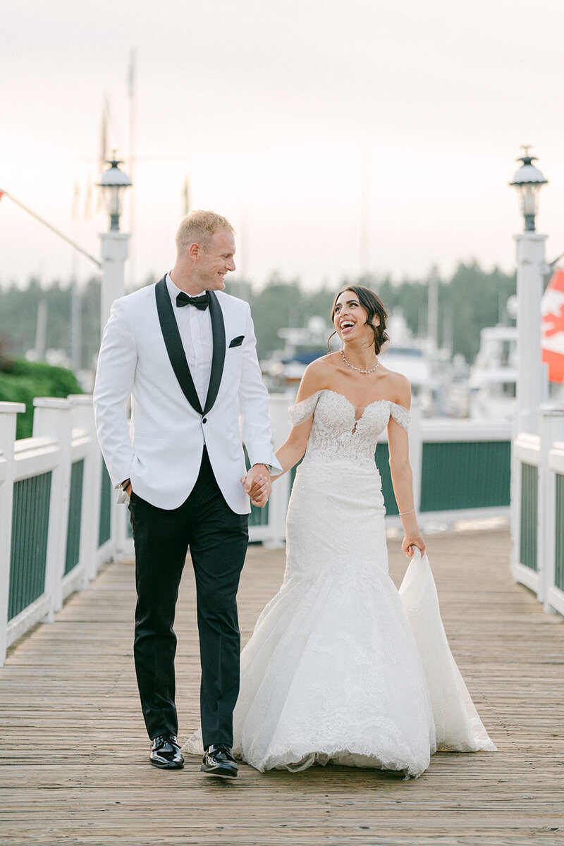 a bride and groom walking on a pier smiling and laughing at each other 