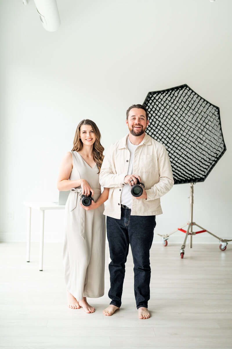 Kala, who looks like she is shooting a picture of you with her camera, is standing in front of Reg who is also holding a camera.