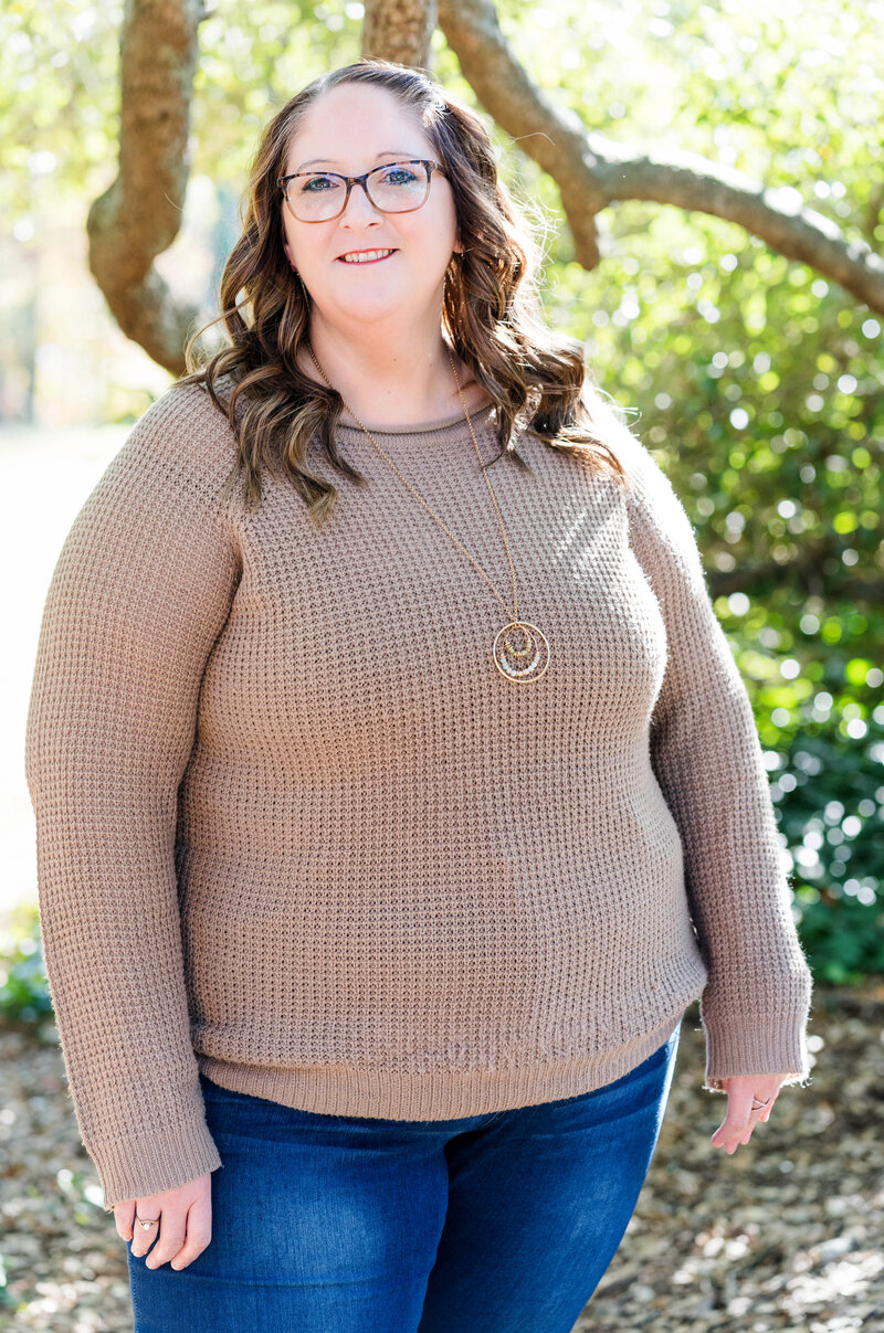 Woman in a tan sweater and jeans standing outdoors surrounded by large twisted tree branches and sunlight filtering through the leaves.