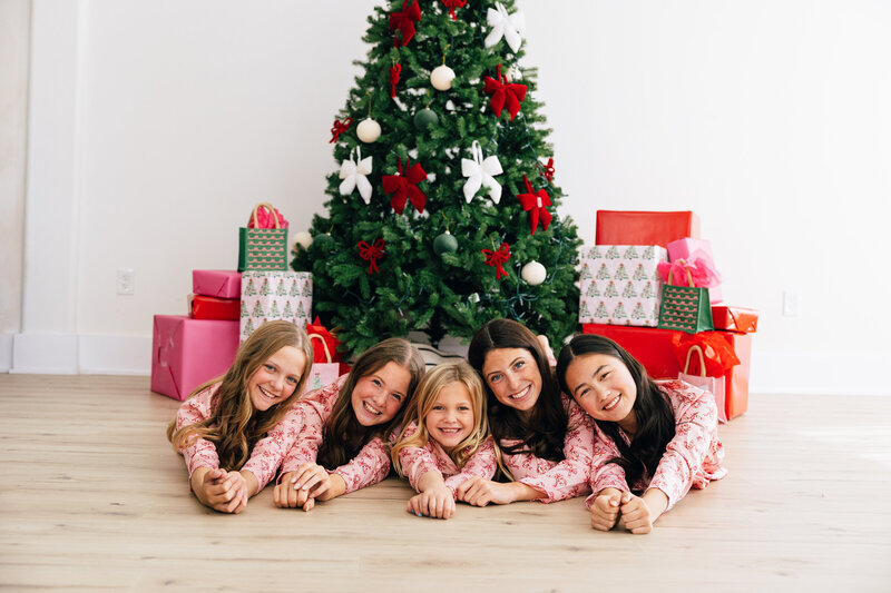 Five sisters laying on stomachs in front of a christmas tree in matching christmas pajamas.