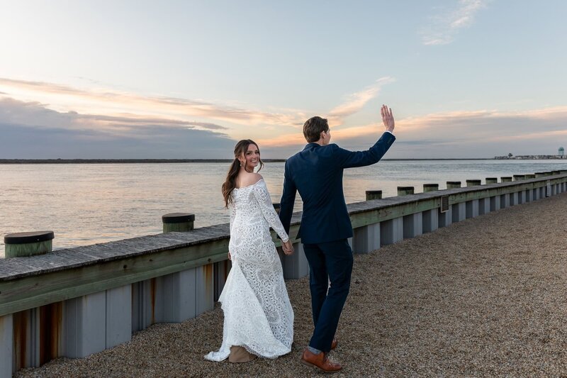 Along the water’s edge at the Jersey Shore, a bride glances back with a joyful smile while holding hands with her new husband. Dressed in a long-sleeved lace gown, she walks the gravel path by the dock as the sunset reflects on the bay. With soft pink clouds overhead, the whole moment feels coastal, romantic, and easy.