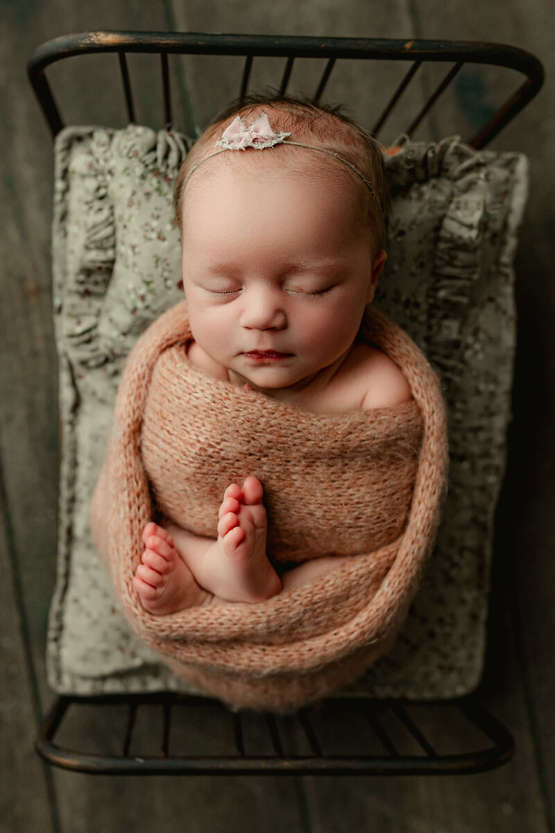 newborn baby girl wrapped in a soft peach wrap with her toes poking out and a tiny pink bow on a newborn bed during newborn session in harrisburg pa